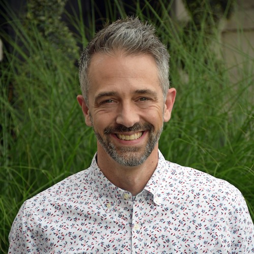 Jason Brommet smiles in front of greenery. He has short grey hair with facial hair and is wearing a button-down shirt.