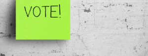 Green sticky note with the word vote on it against white concrete wall