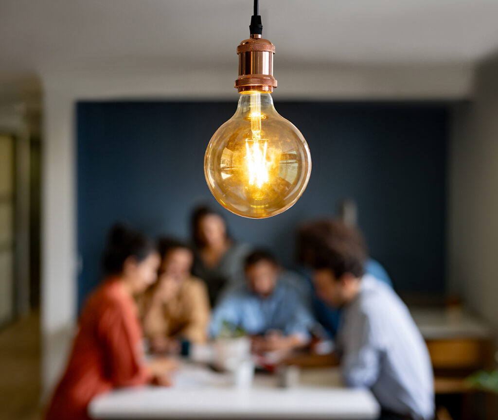 A group gathers around a table, a lightbulb hangs in front of the image of the group to represent ideas and brainstorming.
