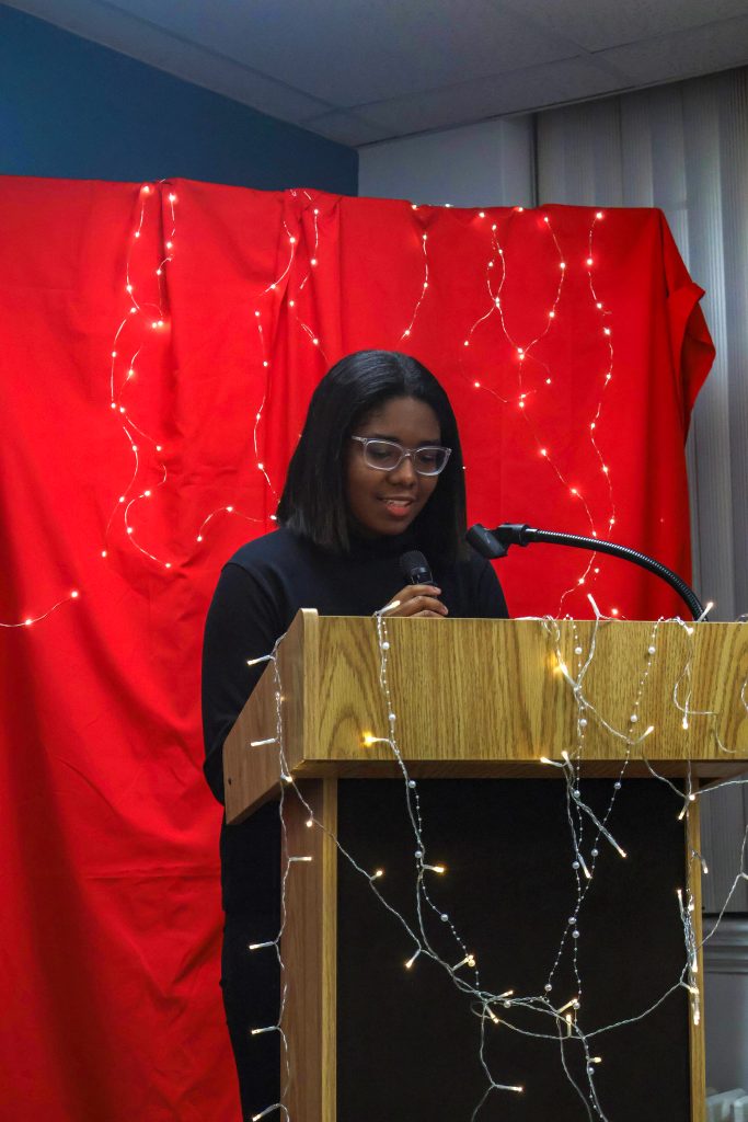 Catherine standing behind the podium and holding a microphone, reading her piece and smiling.