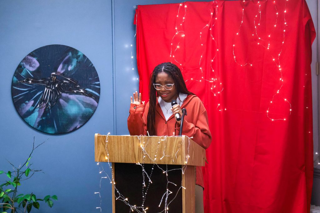 A student holding the microphone in one hand with their other hand up as they read and perform their piece standing behind the podium.