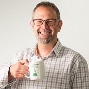 Michal Kasprzak smiles at the camera holding a coffee mug. He has grey hair, a beard and wears glasses. He is wearing a checkered shirt.