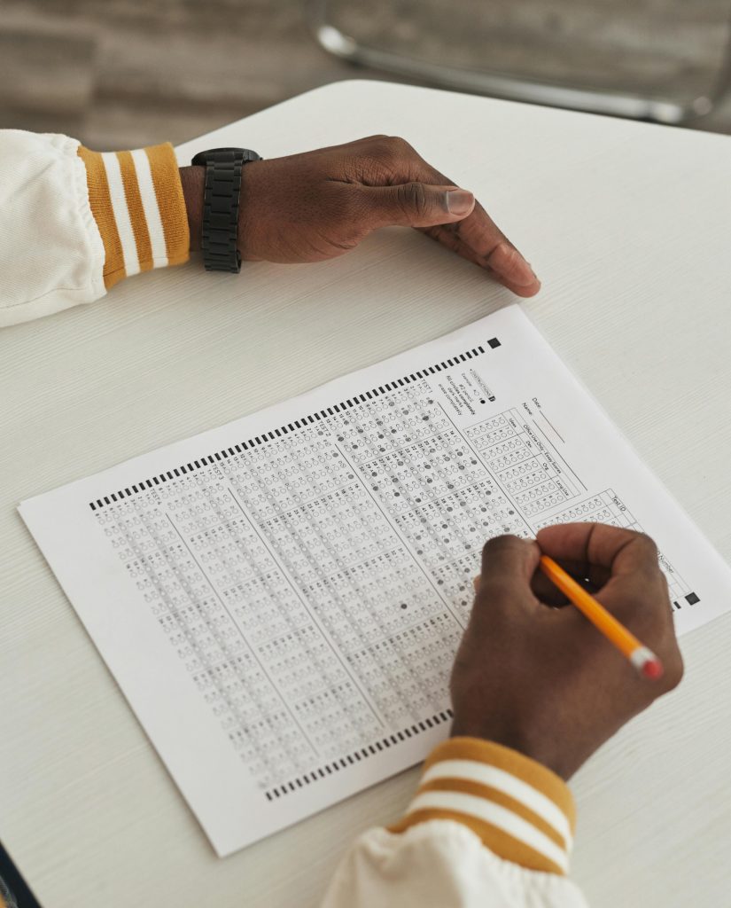 A close up view of a person's hands on a desk marking multiple choice answers on an exam page with a pencil.