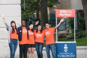 Five AskMe Anything student ambassadors standing together with their arms up, beside an AskMe booth in front of the front campus entrance.