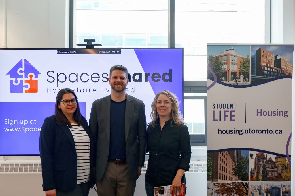 Three people standing together in front of a digital screen that says SpacesShared and a sign that says Student Life Housing.