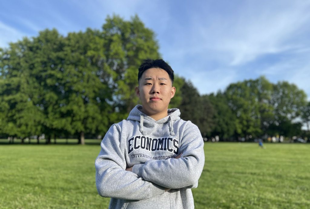 Yu wearing an Economics, University of Toronto hoodie, standing with his arms crossed in an open field.