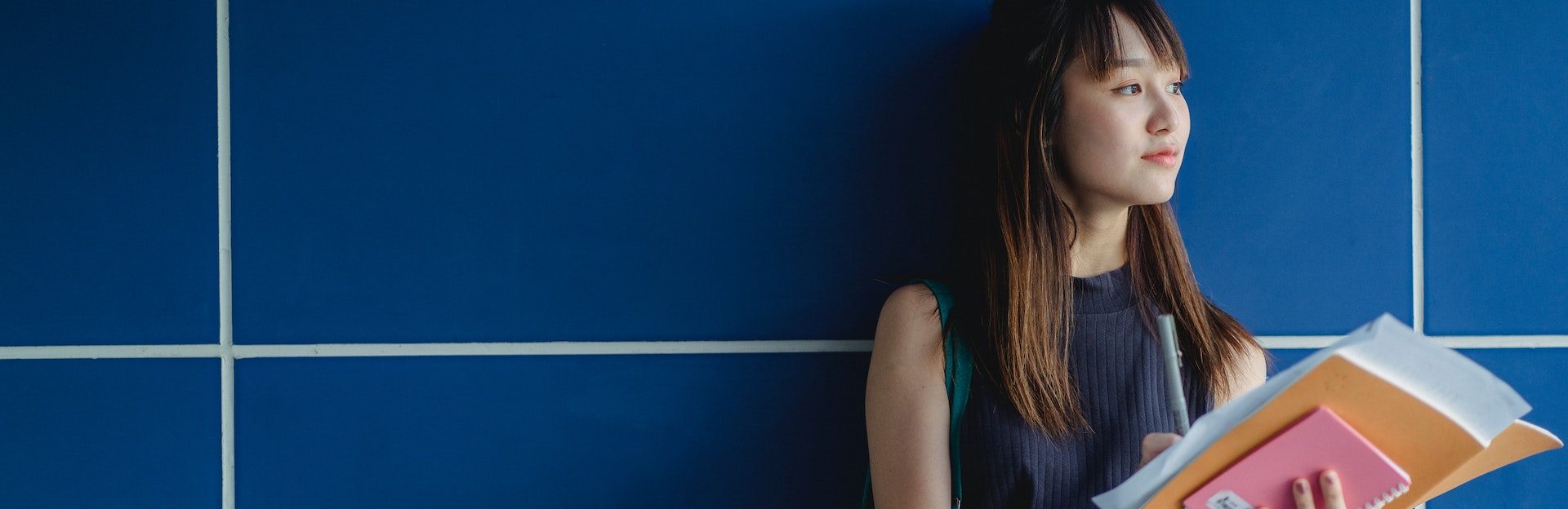 Young woman standing against blue tiled wall holding school books