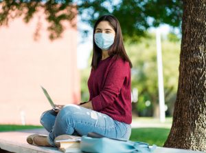 Girl sits outside wearing a red shirt and a mask, smiling over laptop