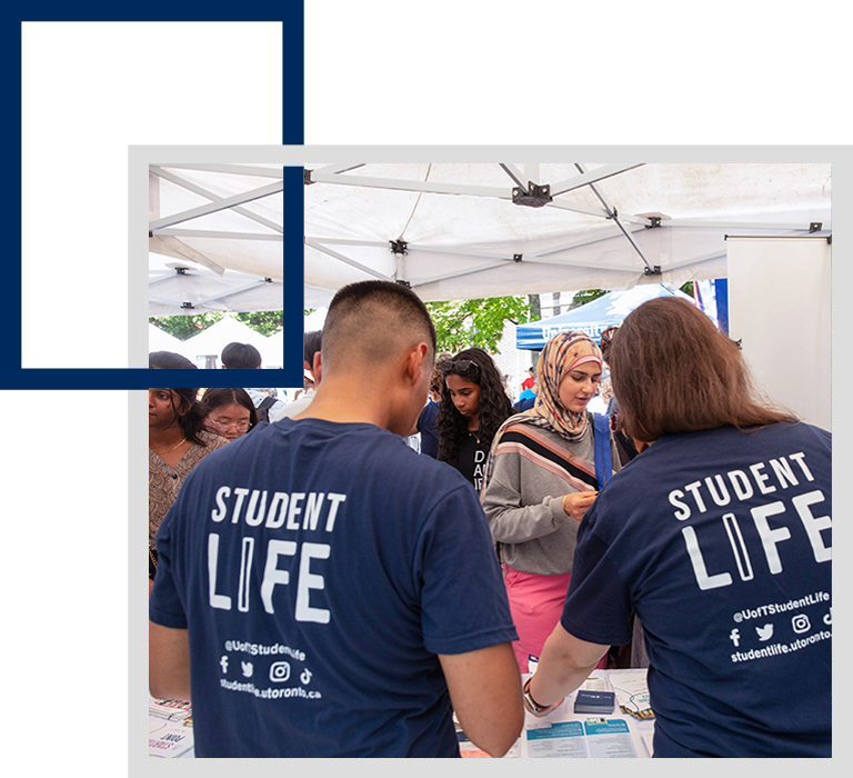 Two people from the Student Life staff team, wearing Student Life t-shirts are seen from behind a booth serving a group of students.