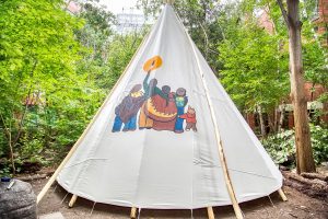 Artwork on tipi: six figures resembling a family, facing forward, embracing shoulder to shoulder. Figure in the middle holds up an eagle feather to the sun.