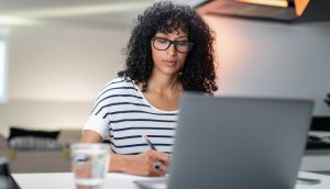 Woman with curly hair and glasses sitting in front of laptop taking notes